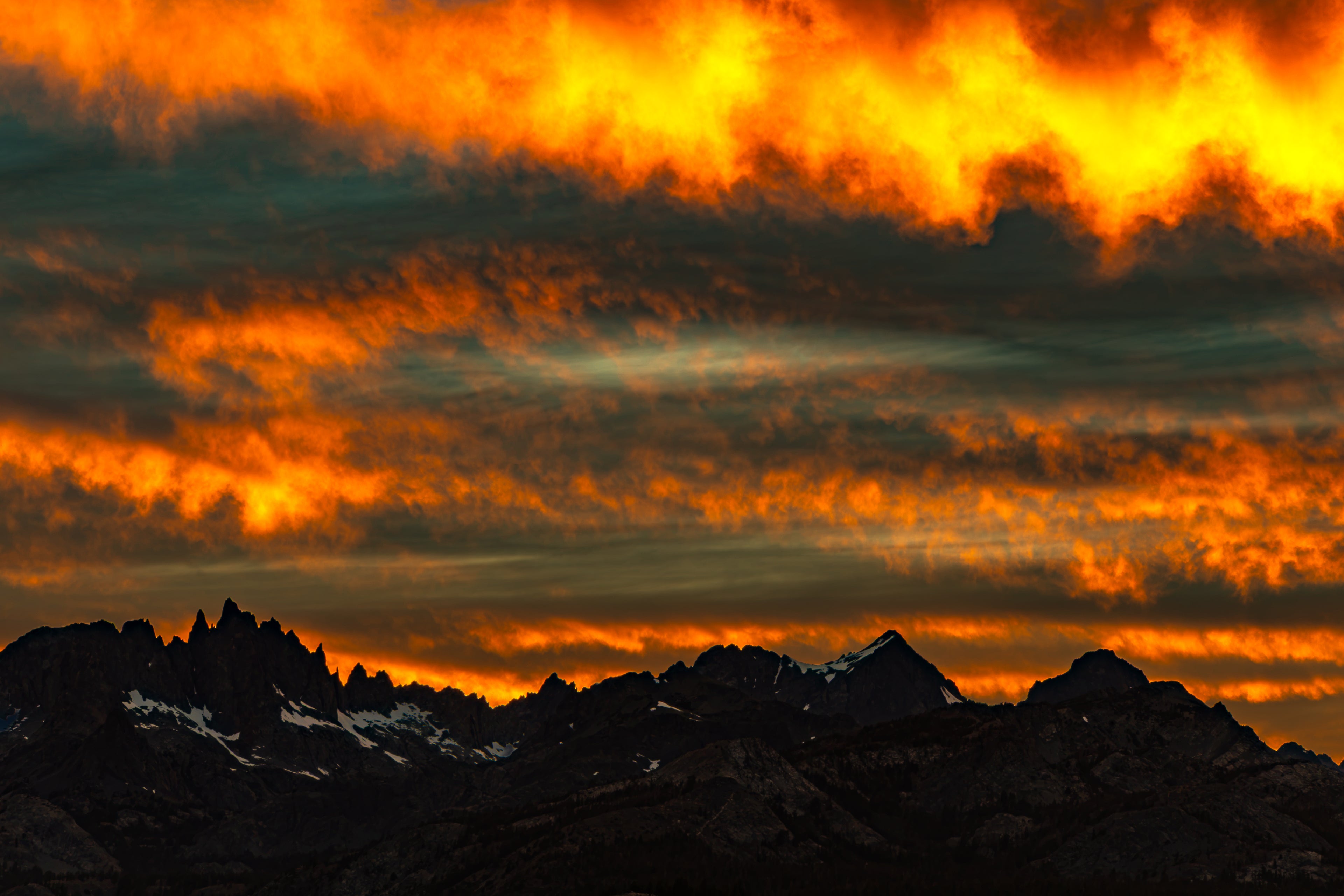 A dramatic mountain landscape at sunset or sunrise, with dark jagged peaks in silhouette against a sky filled with glowing orange, yellow, and brown clouds.
