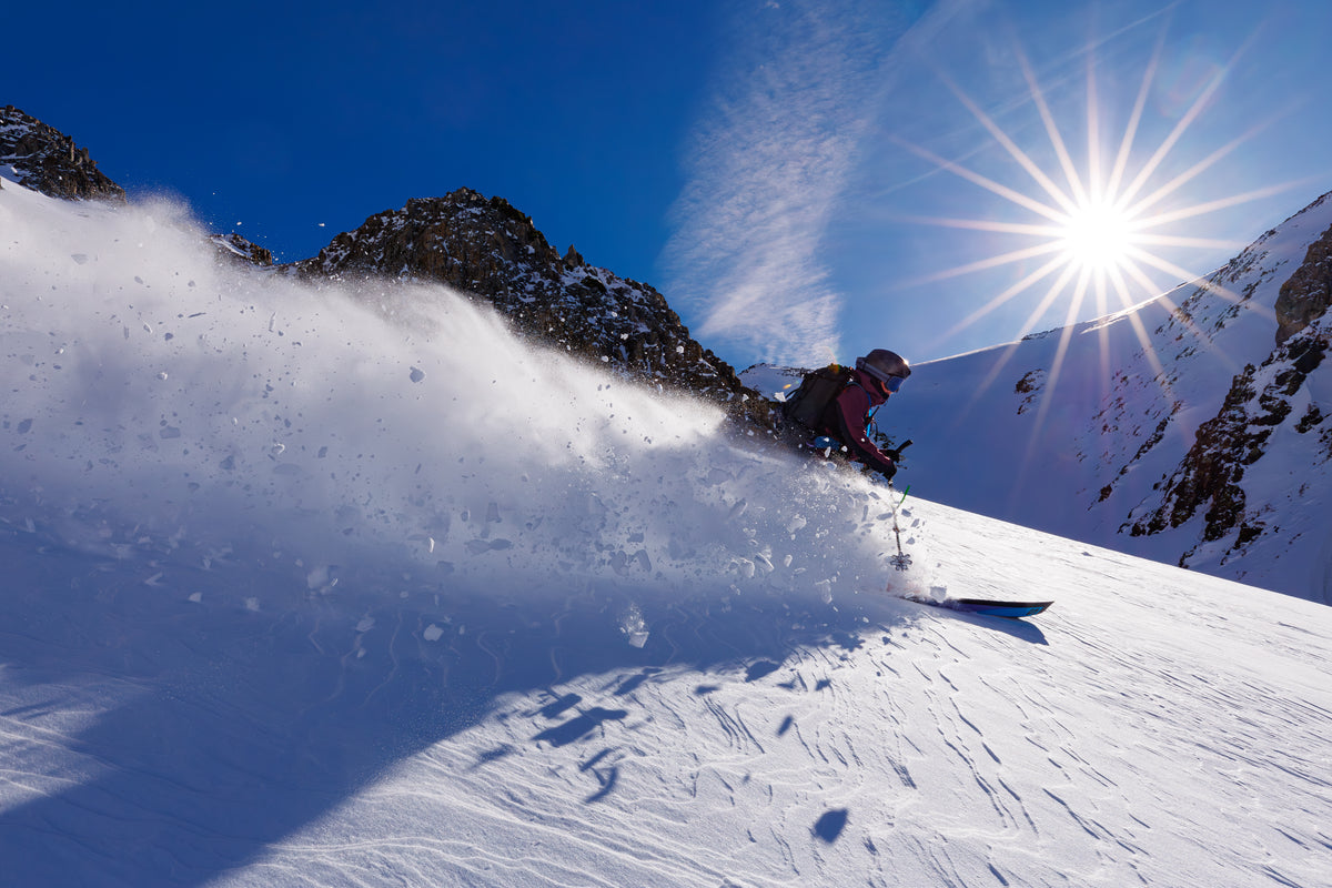 A skier carving through deep powder snow on a sunny mountain slope, with a large spray of snow kicked up behind them. Jagged rocky peaks rise in the background under a bright blue sky with the sun shining in the upper right.